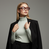 Woman wearing a light green turtleneck sweater, bordeaux glasses and black blazer against a gray background