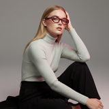 Woman wearing a light gray turtleneck sweater, red frames and dark pants against a neutral background