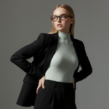 Woman wearing a black blazer, muse cateye frame and light green turtleneck against a gray background